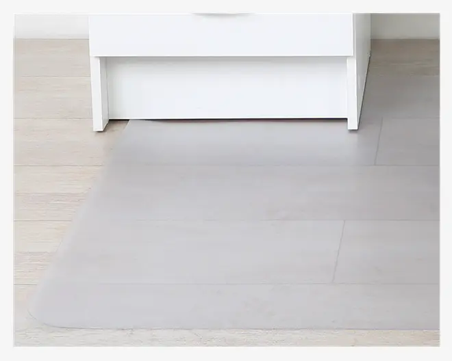 En gennemsigtig, rektangulær gulvmåtte med afrundede hjørner placeret på et lyst trægulv. Måtten ligger foran et hvidt møbel og beskytter gulvet, samtidig med at gulvets naturlige udseende stadig kan ses.