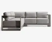 Moderne L-formet sofa med en robust, vinklet metalramme i mat finish. Sofaen har tykke, lysegrå hynder på både sæde og ryg, hvilket giver en blød og komfortabel siddeoplevelse. Armlænene er brede og firkantede, hvilket matcher rammestrukturen og det moderne design. Betrækket ser ud til at være lavet af et slidstærkt, vævet stof, velegnet til både indendørs og udendørs brug.