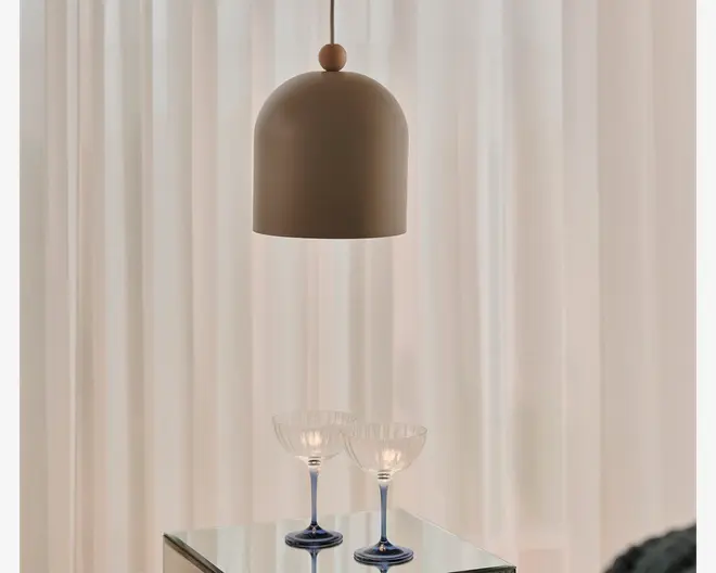 En mat brun kuppelformet pendellampe med en glat, afrundet top og en kort ophængningsledning, vist over et bord med glasplade foran bløde cremefarvede gardiner.