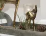 En rustik træbakke i grøn-grå nuancer rummer en lille bronzefarvet hjortefigur, en fyrregren og en kogle, med et sløret lys og guldfarvet dekoration i baggrunden.