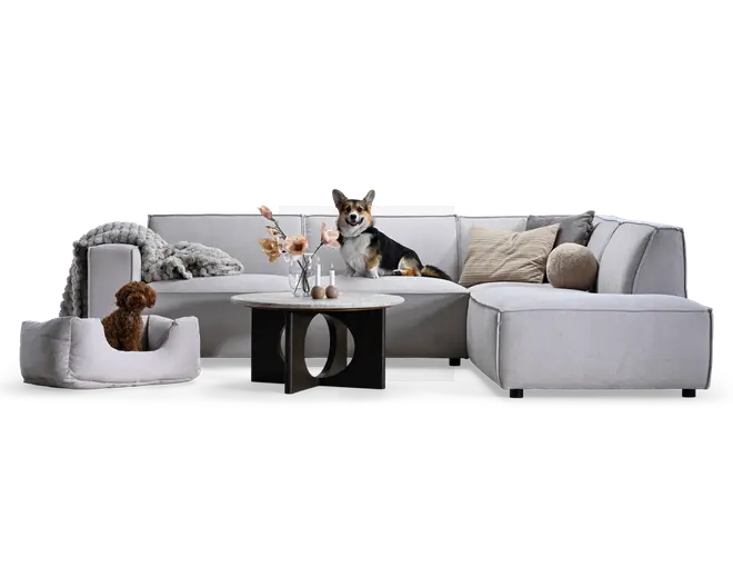 Lysegrå polstret hjørnesofa med chaiselong på højre side, enkle firkantede hynder, lave sorte ben og neutrale pyntepuder, stylet med et rundt sofabord, vase med blomster, stearinlys og bløde tæpper; vist med to hunde for størrelsesfornemmelse.