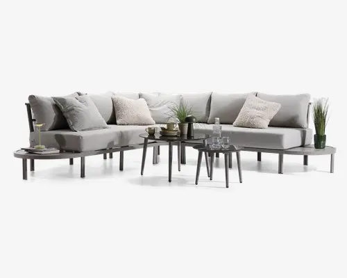 Moderne L-formet sofa med lysegråt betræk og slanke, lige ben. Sædet har bløde, aftagelige hynder og er pyntet med forskellige puder i neutrale nuancer og teksturer. Foran sofaen står to runde, minimalistiske sofaborde med dekorationer som potteplanter, en vandflaske med glas og en kop. Udtrykket er moderne og indbydende, velegnet til stue eller lounge.