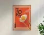 Indrammet plakat med en kraftig orange baggrund, der viser et stiliseret spejlæg, som holdes af et par træspisepinde. Ordet 'TOKYO' står med store, mørkeblå bogstaver, hvor 'O'et delvist overlapper ægget. Dekorativ damp og japanske tegn tilføjer visuel interesse. Rammen er af lyst træ, og plakaten har et moderne, minimalistisk design.