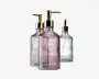 Tre elegante pumpeflasker i glas med lodret rillet struktur og præget 'SOAP'-mærke på forsiden. Hver flaske har en diskret farvetone: én er lyserød, én er klar, og én er lysegrøn. Alle har blanke guldfarvede pumpetoppe, hvilket giver et stilfuldt og luksuriøst udtryk.