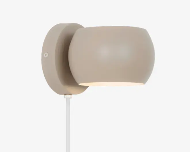 Væghængt lampe med en glat, rund skærm i mat beige og matchende cirkulær base. Lampen har et minimalistisk design med en åbning, der vender nedad for fokuseret belysning. En hvid ledning går ud fra bunden af basen.