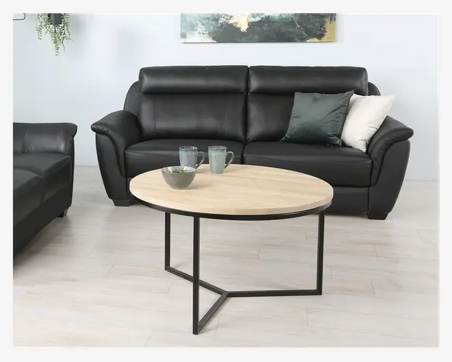 Rundt sofabord med lys træbordplade og sort metalstel med moderne, geometrisk base. Bordet er dekoreret med to keramikkrus og en lille skål, placeret i en moderne stue.