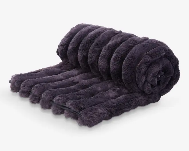 Blød, mørkegrå plaid med imiteret pels og ribbet, stribet mønster. Materialet ser tykt og blødt ud, hvilket giver et luksuriøst og hyggeligt udtryk. Plaiden er delvist rullet sammen, hvilket fremhæver dens fyldige og voluminøse struktur.