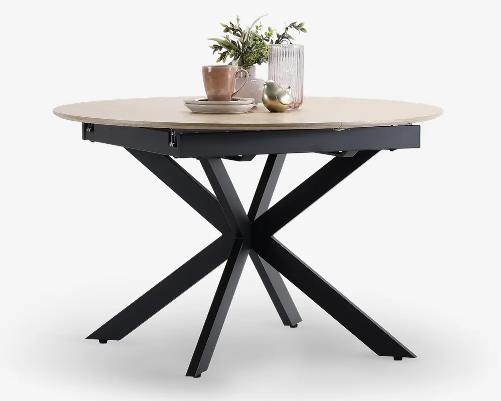 Et rundt bord med en lys træbordplade og en markant sort metalbase med fire skråtstillede ben, der mødes i midten. Bordpladen har en glat overflade og let skrå kanter. På bordet står dekorative genstande, herunder et lyserødt krus, stabelde tallerkener, en rillet glasvase med grønne blade og en metallisk pyntegenstand.