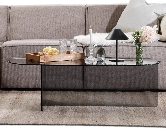 Rektangulært sofabord lavet af røgfarvet glas med afrundede hjørner og et sømløst, buet design. Bordet har et moderne, minimalistisk udtryk med en glat, reflekterende overflade. Det er dekoreret med en træbakke med snacks og glas, en sort bordlampe med konisk skærm, et hvidt stearinlys i en glasstage, dekorative glasgenstande og en grøn vase med lyserøde blomster. Bordet står på et neutralt tæppe foran en beige hjørnesofa med bløde puder.