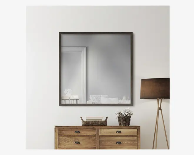 Et stort, kvadratisk vægspejl med en tynd, mørk træramme. Spejlet er monteret over en kommode i træ med fire skuffer og sorte metalhåndtag. Ovenpå kommoden står to flettede kurve, den ene med en lille stak bøger og den anden med en potteplante med hvide blomster. Til højre for kommoden står en høj gulvlampe med trefod og mørkebrun stofskærm. Indretningen er minimalistisk og moderne med neutrale farver og naturlige materialer.