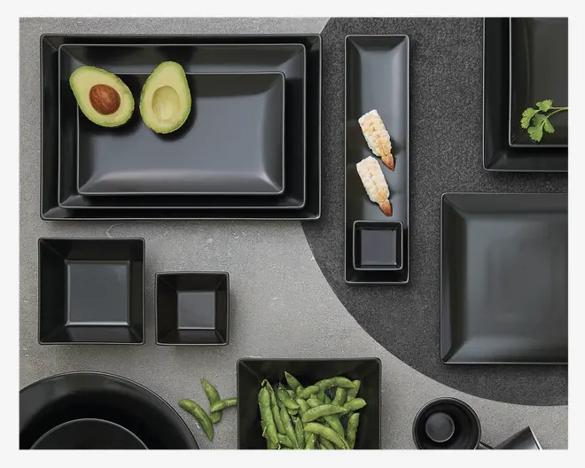 Et sæt mat sorte tallerkener og skåle i forskellige former og størrelser, herunder rektangulære, firkantede og runde. Produkterne har rene, moderne linjer og en glat overflade, hvilket passer til et moderne borddækning. Nogle af delene er vist med mad, såsom halve avocadoer, rejer og edamame, hvilket viser sættets alsidighed til servering af forskellige typer retter.