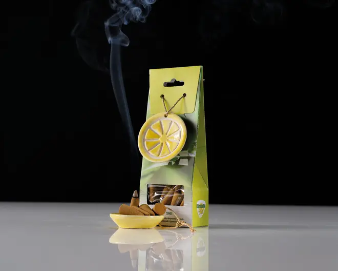 Billedet viser et sæt røgelseskegler med en citroninspireret keramisk holder og et matchende ornament formet som en citronskive. Røgelseskeglerne er placeret i holderen, hvoraf én kegle brænder og udsender en tydelig røgstribe. Emballagen står oprejst og har et vindue, der viser keglerne indeni, og citronskive-ornamentet er fastgjort på forsiden. Det samlede design fremhæver et frisk citrus-tema med gule og grønne farver.
