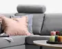 Grå polstret hjørnesofa med rund cylinderformet nakkestøtte, dybe sædehynder og to dekorative pyntepuder i pudderrosa og lyseblå.