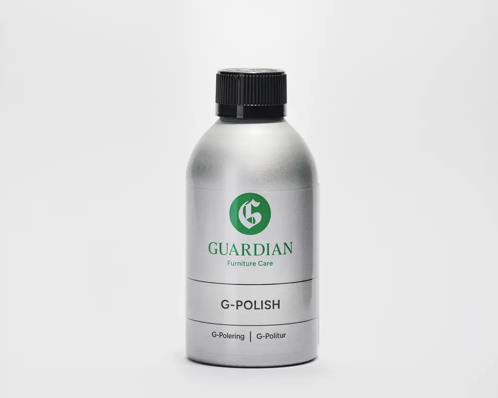 En sølvfarvet flaske med sort skruelåg, der har et grønt 'G'-logo og mærket 'Guardian Furniture Care' på forsiden. Etiketten viser også produktnavnet 'G-Polish' med fed skrift samt yderligere tekst i mindre skrift med 'G-Polering' og 'G-Politur.' Flasken har et slankt, moderne design og ser ud til at være lavet af metal eller metallisk plast.