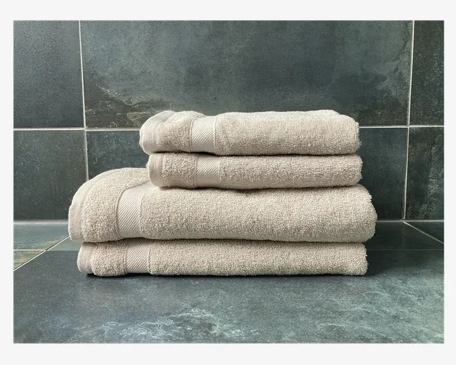 Et sæt på fire beige håndklæder i forskellige størrelser, pænt stablet fra det største nederst til det mindste øverst. Hvert håndklæde har en diskret tekstureret overflade og et dekorativt bånd nær den ene kant, hvilket giver et blødt og fyldigt udtryk. Håndklæderne er præsenteret mod en mørk flisebaggrund, hvilket fremhæver deres neutrale farve og rene finish.