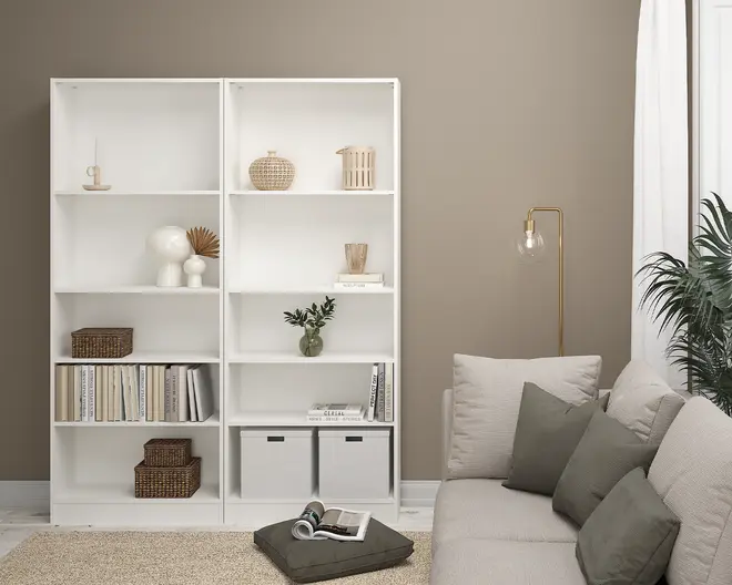 Høj, hvid reol med flere åbne rum, der indeholder en blanding af dekorative genstande som flettede kurve, keramiske vaser, bøger og to stofopbevaringskasser nederst. Hylderne er jævnt fordelt, og det samlede design er minimalistisk og moderne, velegnet til organisering af bøger og udstilling af pynt.