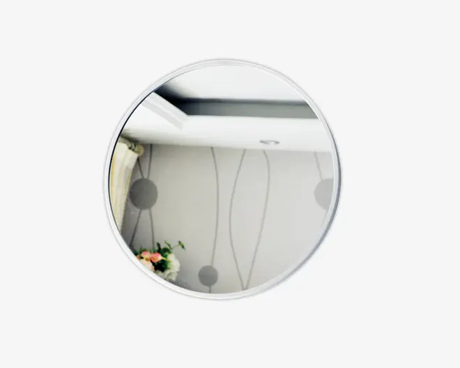 Et rundt spejl med en tynd, sølvfarvet metalramme. Spejlet har en klar refleksion, hvor man kan se en del af et rum med mønstret tapet, et loft med indbygget belysning og en lille buket blomster i hjørnet. Designet er moderne og minimalistisk.