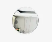 Et rundt spejl med en tynd, sølvfarvet metalramme. Spejlet har en klar refleksion, hvor man kan se en del af et rum med mønstret tapet, et loft med indbygget belysning og en lille buket blomster i hjørnet. Designet er moderne og minimalistisk.