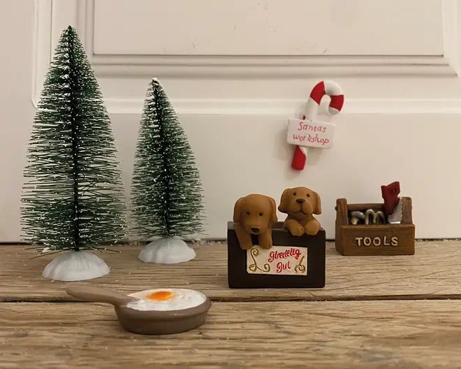 Billedet viser en samling af små juledekorationer arrangeret på en træoverflade. Der er to grønne juletræer lavet af flaskerensere med hvide fodder, en lille brun kasse med to brune hundehvalpefigurer og et skilt, hvor der står 'glædelig jul', en lille værktøjskasse med mærket 'TOOLS', der indeholder miniatureværktøj, samt en lille stegepande med et spejlæg. I baggrunden er der en slikstok med et skilt, hvor der står 'Santa's workshop', som er sat op på væggen.