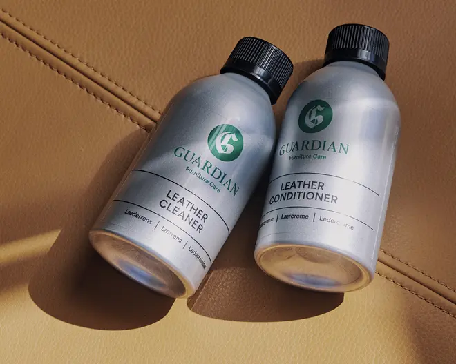 To sølvfarvede flasker med sorte skruelåg, mærket 'Guardian Furniture Care.' Den ene flaske er mærket 'Leather Cleaner' og den anden 'Leather Conditioner.' Begge flasker har et minimalistisk design med grøn og sort tekst og ligger på en lysebrun læderoverflade med synlige syninger. Flaskerne ser robuste ud og er nemme at håndtere.
