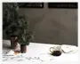 Billedet viser en hvid dug med broderede grangrene og kogler. På bordet står to kunstige grantræer i rustikke potter med bark, et klart glas krus med guldfarvet hank fyldt med en mørk drik, samt en klar glas-tallerken med en sammenfoldet serviet, der har et lille broderet grangren-motiv. Hele opstillingen har et naturligt, vinter- eller juleinspireret tema.
