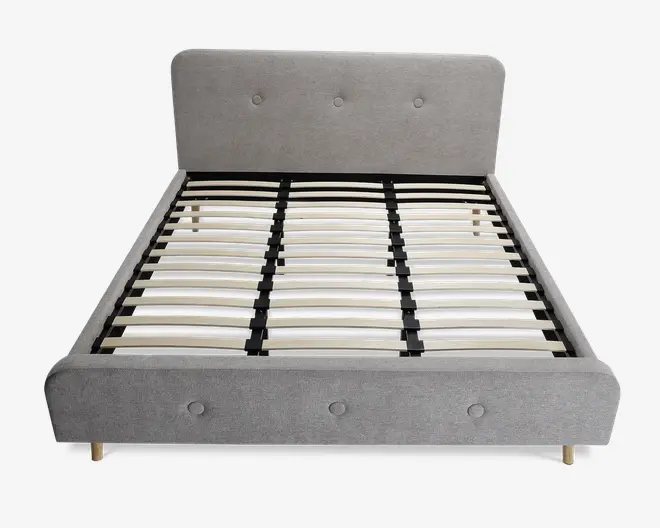 En moderne sengeramme med lysegråt polstret hovedgærde og fodgærde, begge med knapdetaljer. Rammen har lameller af træ til at støtte madrassen og står på korte, lyse træben. Designet er minimalistisk og nutidigt med rene linjer og en blød tekstiloverflade.
