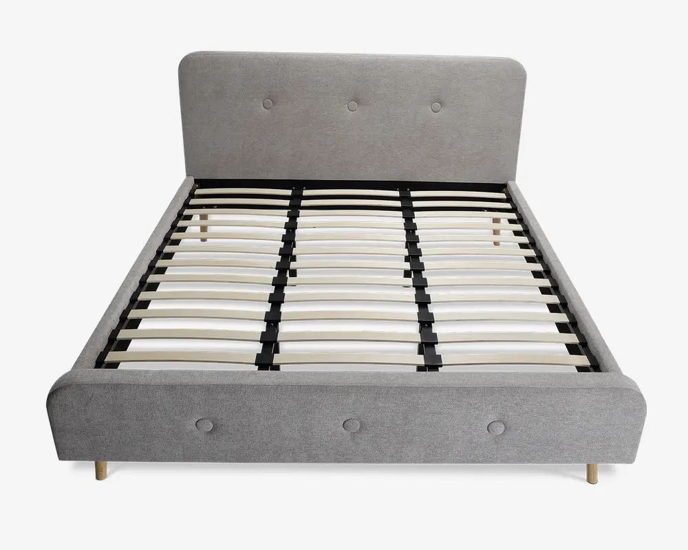 En moderne sengeramme med lysegråt polstret hovedgærde og fodgærde, begge med knapdetaljer. Rammen har lameller af træ til at støtte madrassen og står på korte, lyse træben. Designet er minimalistisk og nutidigt med rene linjer og en blød tekstiloverflade.