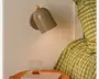 Mat beige væglampe med afrundet bagplade, buet kegleformet skærm og lille træknop, vist over et natbord med et klart glas og grøn- og lyserødstribet sengetøj.
