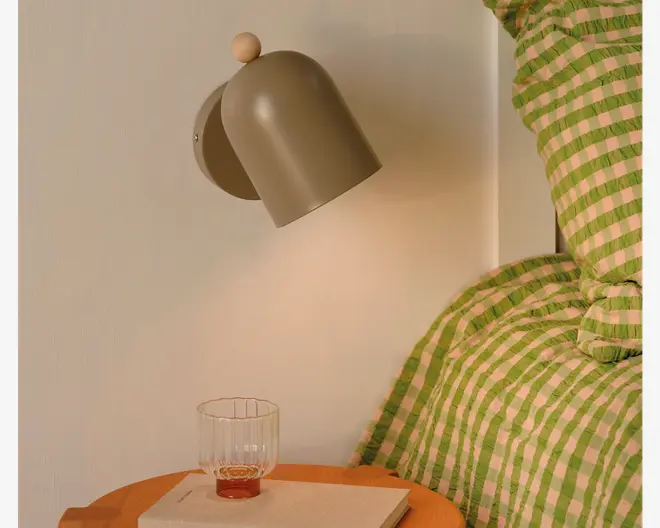 Mat beige væglampe med afrundet bagplade, buet kegleformet skærm og lille træknop, vist over et natbord med et klart glas og grøn- og lyserødstribet sengetøj.