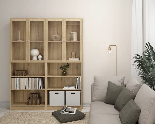 Høj reolenhed i lyst træ med naturlig finish, bestående af to sektioner med hver fire hylder. De øverste hylder har glaslåger, mens de nederste er åbne. Hylderne er dekoreret med forskellige genstande som vaser, kurve og bøger. To hvide opbevaringskasser står på den nederste højre hylde. Reolen står på et lyst gulv ved siden af en beige sofa med grå og grønne puder.