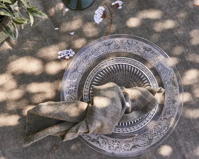En klar glasstallerken med en indviklet, dekorativ kant og et tekstureret, strålende mønster i midten. Tallerkenen ligger på en neutralfarvet, tekstureret dug med diskret blomsterbroderi. En linnedserviet i en afdæmpet, jordfarvet tone er pænt foldet og holdt sammen af en enkel servietring, som ligger hen over tallerkenen. Plettet sollys skaber bløde, naturlige skygger på borddækningen.