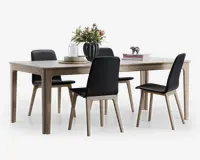 Rektangulært spisebord med lys træfinish og fire matchende stole. Stolene har sorte polstrede sæder og rygge med ben i lyst træ. Bordet er dekoreret med en grøn vase med blomster, stakke af bøger samt et sæt tallerkener og stearinlys, hvilket giver et moderne og elegant udtryk.