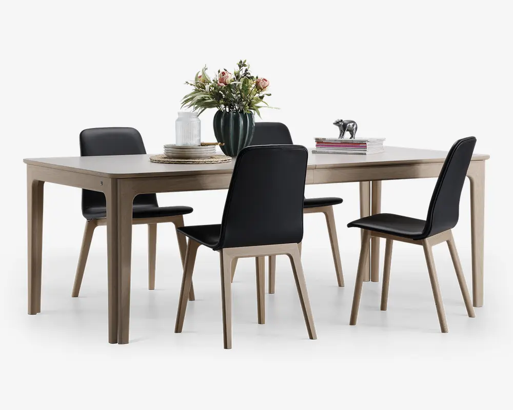 Rektangulært spisebord med lys træfinish og fire matchende stole. Stolene har sorte polstrede sæder og rygge med ben i lyst træ. Bordet er dekoreret med en grøn vase med blomster, stakke af bøger samt et sæt tallerkener og stearinlys, hvilket giver et moderne og elegant udtryk.