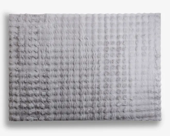 Rektangulært måtte med en blød, plysset tekstur og et diskret gittermønster. Overfladen fremstår luftig og tyk, hvilket giver en komfortabel og hyggelig fornemmelse. Farven er en lys, neutral grå, hvilket gør den alsidig til forskellige indretninger. Kanterne er lige og pænt afsluttede.