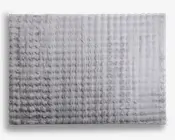 Rektangulært måtte med en blød, plysset tekstur og et diskret gittermønster. Overfladen fremstår luftig og tyk, hvilket giver en komfortabel og hyggelig fornemmelse. Farven er en lys, neutral grå, hvilket gør den alsidig til forskellige indretninger. Kanterne er lige og pænt afsluttede.