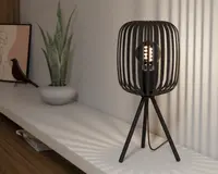 Moderne bordlampe med sort metalskærm i bur-design og tre slanke ben i tripod-stil. Pæren er synlig gennem de åbne lameller, hvilket skaber et dekorativt lysmønster på væggen. Lampen har et minimalistisk, nutidigt design og en sort ledning.