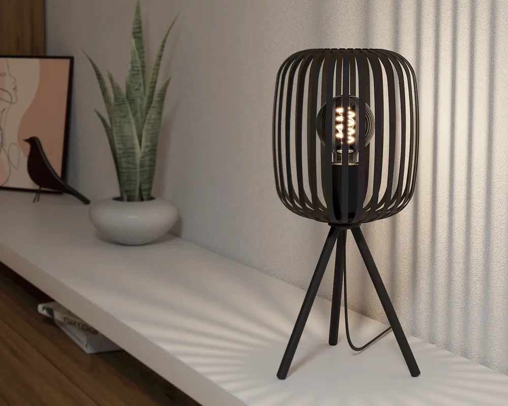 Moderne bordlampe med sort metalskærm i bur-design og tre slanke ben i tripod-stil. Pæren er synlig gennem de åbne lameller, hvilket skaber et dekorativt lysmønster på væggen. Lampen har et minimalistisk, nutidigt design og en sort ledning.