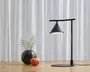 En moderne bordlampe med mat sort finish, der har en konisk skærm ophængt fra en vandret arm, som er fastgjort til en lodret stander. Lampen har en rund base og udsender et blødt, fokuseret lys nedad. Ved siden af lampen står en rund sort vase med fine hvide blomster og en rød granatæble placeret foran, alt sammen på en lys træoverflade med hvide, gennemsigtige gardiner i baggrunden.
