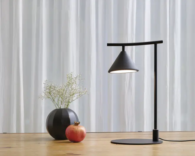 En moderne bordlampe med mat sort finish, der har en konisk skærm ophængt fra en vandret arm, som er fastgjort til en lodret stander. Lampen har en rund base og udsender et blødt, fokuseret lys nedad. Ved siden af lampen står en rund sort vase med fine hvide blomster og en rød granatæble placeret foran, alt sammen på en lys træoverflade med hvide, gennemsigtige gardiner i baggrunden.