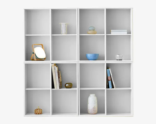 En stor, hvid reol med et gitter af seksten kvadratiske rum. Hylderne er sparsomt dekoreret med forskellige genstande, herunder et lille rundt spejl med træramme, en stribet keramikkop, en blå skål, et glas, et par stablede bøger, nogle dekorative vaser og nogle opretstående bøger. Det overordnede udtryk er minimalistisk og moderne med rene linjer og en neutral farvepalet.