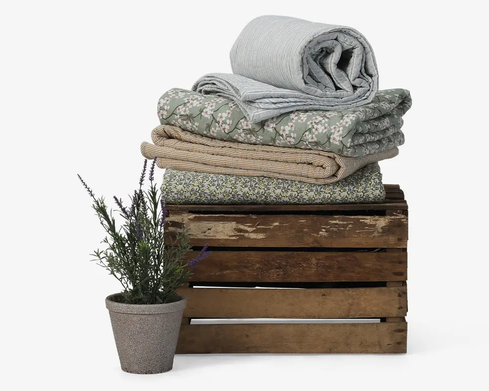En stak pænt sammenfoldede tæpper i forskellige mønstre og farver, herunder lyseblå striber, grøn med hvidt blomstermønster, beige og grøn med små blomster. Tæpperne ligger oven på en rustik trækasse. Foran kassen står en lille potte med lavendelplante med grønne blade og lilla blomster.
