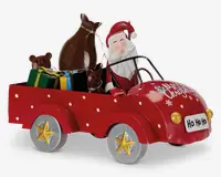 Festlig dekorationsfigur med julemanden, der kører en rød bil med hvide prikker og teksten 'Merry Christmas' på motorhjelmen. Bilen har sølvfarvede hjul med gyldne stjerner og en nummerplade foran med teksten 'Ho Ho Ho'. Bagpå er der farverige indpakkede gaver, en brun sæk og to brune rensdyrfigurer.