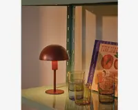 En lille, moderne bordlampe med mat rødbrun finish og kuppelformet skærm står på en hylde. Lampen har et enkelt, minimalistisk design med rund fod og lige stang. Ved siden af lampen står fire farvede glas i nuancer af rav, grøn og blå. Bag glassene står en kogebog med lilla og orange omslag med titlen 'ITALIAN.' Hylden er lysegrøn og lavet af metal.