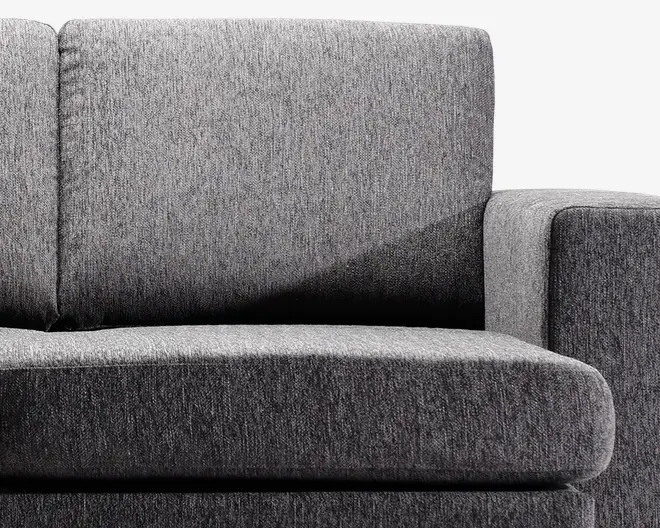 Nærbillede af en moderne sofasektion betrukket med mørkegråt stof. Sæde- og ryghynderne er tykke og bløde, hvilket giver en behagelig siddeoplevelse. Stoffet har en diskret tekstureret vævning, og armlænet er bredt og polstret for ekstra komfort. Designet er minimalistisk med rene linjer.