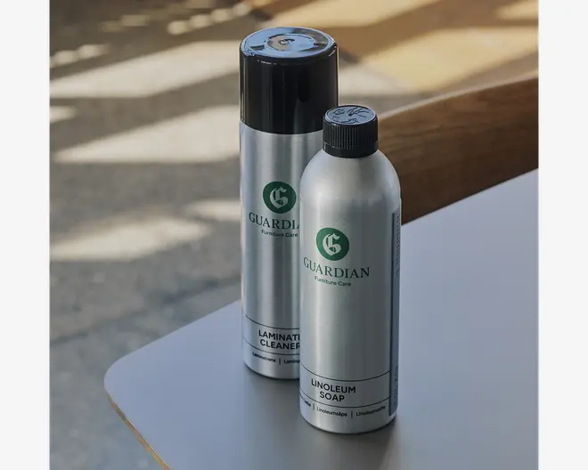 To metalflasker med grøn Guardian-branding, den ene mærket 'Laminate Cleaner' med sort spraylåg og den anden mærket 'Linoleum Soap' med sort skruelåg. Begge flasker har et stilrent, moderne design og tydelig mærkning for nem genkendelse.