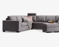 Moderne L-formet hjørnesofa betrukket med lysegråt stof, med brede armlæn og bløde ryghynder. Sofaen har en matchende skammel og et nakkestøtte på ét sæde for ekstra komfort. Den er pyntet med dekorative puder i rosa og hvid samt et tekstureret gråt tæppe, der er lagt over siden. En træbakke med drikkevarer og frugt står på skamlen, hvilket viser dens funktion som sofabord.