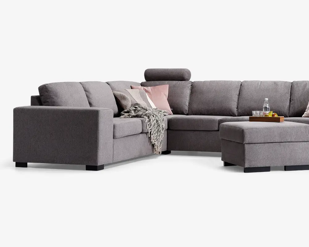 Moderne L-formet hjørnesofa betrukket med lysegråt stof, med brede armlæn og bløde ryghynder. Sofaen har en matchende skammel og et nakkestøtte på ét sæde for ekstra komfort. Den er pyntet med dekorative puder i rosa og hvid samt et tekstureret gråt tæppe, der er lagt over siden. En træbakke med drikkevarer og frugt står på skamlen, hvilket viser dens funktion som sofabord.