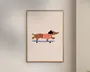 Minimalistisk illustration af en brun gravhund iført en rød- og hvidstribet sweater, der kører på et blåt skateboard. Baggrunden er ensfarvet lys beige, og kunstværket er indrammet i en enkel træramme.