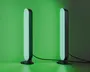 Et par slanke, lodrette LED-lysstænger med afrundede kanter, der udsender et blødt grønt skær. Hver lysstang er monteret på en lille, rund fod og forbundet med en synlig sort strømkabel. Lysene står på en flad overflade op ad en væg og skaber en stemningsfuld belysning.