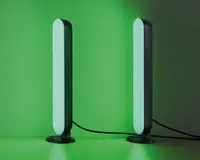 Et par slanke, lodrette LED-lysstænger med afrundede kanter, der udsender et blødt grønt skær. Hver lysstang er monteret på en lille, rund fod og forbundet med en synlig sort strømkabel. Lysene står på en flad overflade op ad en væg og skaber en stemningsfuld belysning.